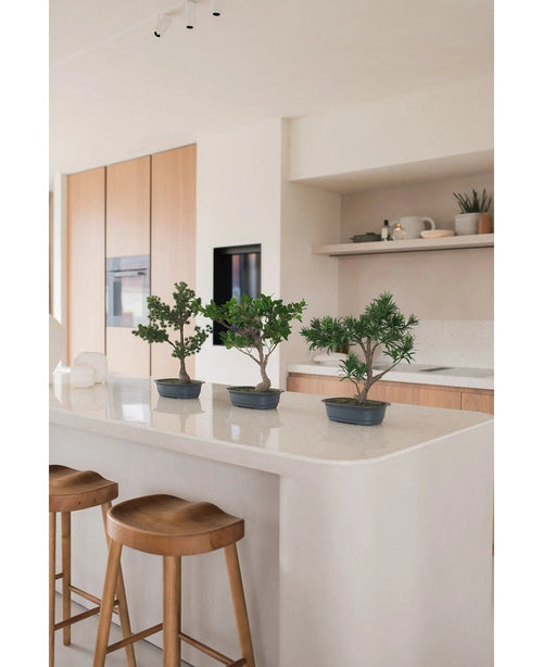 Una pianta artificiale Bonsai Ficus da 40 cm si trova su un'isola da cucina bianca con due sgabelli in legno, circondata da mobili in legno chiaro, piani di lavoro bianchi e scaffali dall'arredamento minimal.