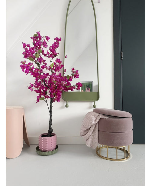 Un interno moderno con la Bougainvillea artificiale viola da 125 cm in un vaso decorativo, uno specchio alto con cornice verde, un poggiapiedi rosa con base dorata e coperta rosa e una porta scura contro un muro bianco frizzante.