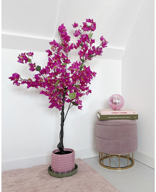 La bouganville artificiale viola da 125 cm con vivaci fiori viola si trova accanto a uno sgabello in velluto rosa con libri impilati sopra e una palla di vetro rosa in una stanza minimale e moderna con pareti bianche.