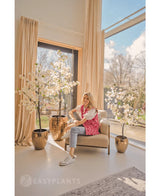 Een vrouw lacht op een beige fauteuil bij grote ramen met beige gordijnen, omringd door drie Kunstplant Bougainvillea 150 cm wit in een lichte, moderne leefruimte vol natuurlijk licht.