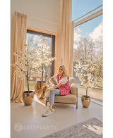 Een vrouw in een roze vest met patroon en spijkerbroek zit op een beige fauteuil bij grote ramen met crèmekleurige gordijnen, omringd door drie Kunstplant Bougainvillea van 180 cm wit in glanzend gouden plantenbakken. Zonlicht vult de moderne, luchtige kamer.