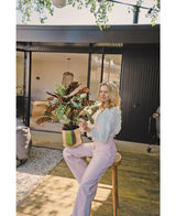 Een blonde vrouw in een witte blouse en lichtpaarse broek zit buiten op een krukje met de Kunstplant Calathea Makoyana UV 75 cm rode potplant, op een houten vlonder voor een modern huis met glazen deuren.