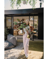 Een vrouw staat op een houten dek voor een modern huis, glimlacht en houdt twee Kunstplant Calathea Makoyana UV 75 cm rode potplanten vast. Ze draagt een lichtroze broek en een wit topje. Buiten zitplaatsen en groen op de achtergrond.
