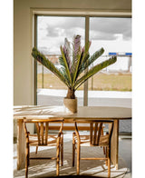 Een houten eettafel met twee bijpassende stoelen staat bij een groot raam. Op de tafel staat een Kunstplant Cycus Palm Deluxe 95 cm en het zonlicht stroomt naar binnen en werpt zijn schaduwen op het meubilair en de vloer.