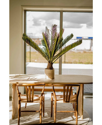 Een houten eettafel met twee bijpassende stoelen staat bij een groot raam. Op de tafel staat een Kunstplant Cycus Palm Deluxe 95 cm en het zonlicht stroomt naar binnen en werpt zijn schaduwen op het meubilair en de vloer.