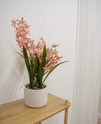 De Kunstplant Cymbidium 58 cm roze, met hoge groene bladeren en roze orchideeachtige bloemen, staat op een lichte houten tafel tegen een effen witte muur en een wit gordijn.