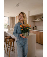 Een vrouw met krullend blond haar, in een denim shirt en spijkerbroek, staat in een moderne keuken met de Kunstplant Daisy UV 31 cm in haar hand, glimlacht en kijkt een beetje opzij.
