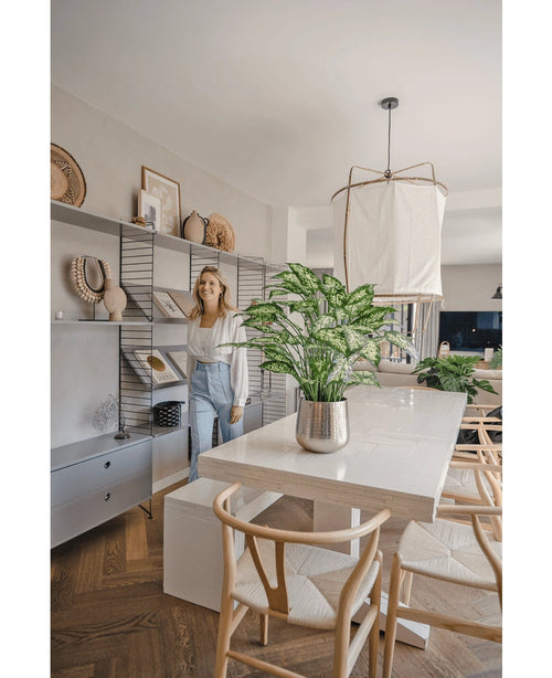 Una donna sorride in una sala da pranzo moderna e luminosa con sedie in legno, un tavolo bianco con la pianta artificiale Dieffenbachia da 75 cm come centrotavola e scaffali aperti decorati con libri e opere d'arte.