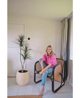 Een vrouw in een felroze topje en blauwe spijkerbroek zit op blote voeten op een moderne zwarte en rieten stoel naast de Kunstplant Dracaena Fragnans 150 cm in een lichte, minimalistische kamer met witte muren en vloer.