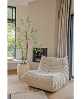 Een gezellige, crèmekleurige loungestoel met textuur staat naast de Kunstplant Enkianthus 210 cm en een moderne open haard in een lichte, minimalistische woonkamer met lichte houten vloeren en grote ramen.