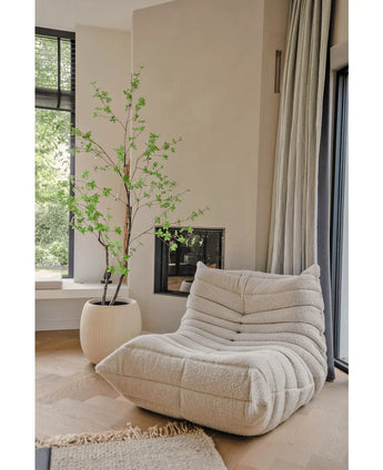 Een gezellige, crèmekleurige loungestoel met textuur staat naast de Kunstplant Enkianthus 210 cm en een moderne open haard in een lichte, minimalistische woonkamer met lichte houten vloeren en grote ramen.