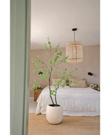 Een minimalistische slaapkamer met beige muren, een wit bed, een natuurlijk geweven tapijt en de Kunstplant Enkianthus 210 cm - een grote potplant met groene bladeren - op de voorgrond. Aan het plafond hangt een geweven hanglamp.