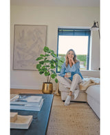 Een vrouw zit op een beige bank in een gezellige, moderne woonkamer, met een mok in de hand en een glimlach. De ruimte wordt gekenmerkt door de Kunstplant Ficus 120 cm in een gouden pot, abstracte kunstwerken en zonlicht dat door blauwe jaloezieën naar binnen schijnt.
