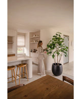 Een vrouw in neutrale kleding staat in een moderne, minimalistische keuken en raakt de bladeren van de Kunstplant Ficus van 210 cm aan. Zonlicht benadrukt de strakke lijnen en aardetinten van de ruimte.