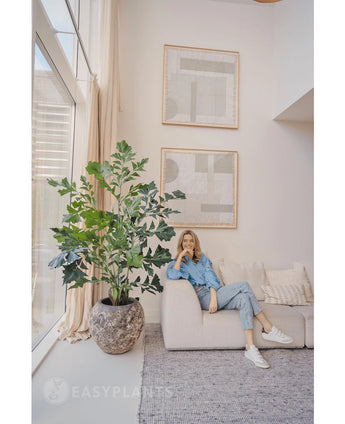 Een vrouw in spijkerstof zit op een lichte bank naast een Kunstplant Fishtail Palm 160 cm in een lichte, moderne woonkamer met minimalistische inrichting en abstracte muurkunst.