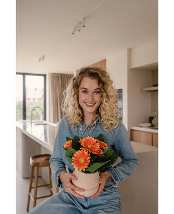 Een glimlachende vrouw met krullend blond haar in spijkerstof zit in een moderne keuken en houdt de Kunstplant Gerbera UV 28 cm oranje vast. Helder natuurlijk licht stroomt door grote ramen achter haar.