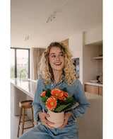 Een glimlachende blonde vrouw in spijkerstof zit in een moderne keuken met de Kunstplant Gerbera UV 28 cm oranje in de hand - een levendige kunstmatige oranje gerberapot. Zonlicht stroomt door een groot raam en verlicht de ruimte.