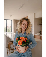 Een vrouw met krullend blond haar in spijkerstof lacht terwijl ze de Kunstplant Gerbera UV 28 cm oranje vasthoudt in een lichte, moderne keuken.