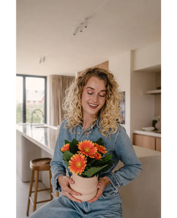 Een vrouw met krullend blond haar in spijkerstof lacht terwijl ze de Kunstplant Gerbera UV 28 cm oranje vasthoudt in een lichte, moderne keuken.
