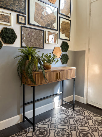 Een hal met gedessineerde vloertegels, een houten consoletafel met daarop de Kunstplant Gouden Wijnstok in Pot 25cm, en een galeriewand met ingelijste kaarten en zeshoekige moskunst op een tweekleurige grijs-witte muur.