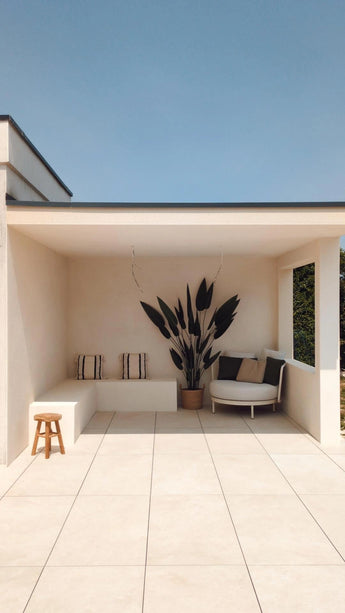 Een moderne, minimalistische patio met crèmekleurige tegels, een ingebouwde bank met gestreepte kussens, een Kunstplant Heliconia 210 cm voor een luxe touch, een klein houten krukje en een ronde stoel met kussens onder een plat dak tegen een helderblauwe hemel.