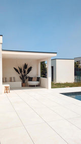 Een modern buitenterras met witte tegelvloeren, schaduwrijke zitplaatsen met een luxe uitstraling, de Kunstplant Heliconia 210 cm als middelpunt, gezellige kussens, een houten kruk en een zwembad op de voorgrond onder een strakblauwe hemel.