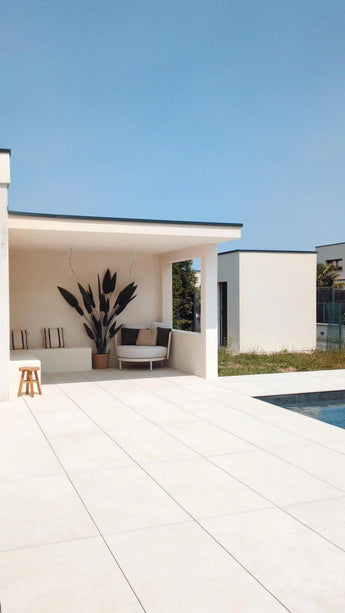 Een modern buitenterras met witte tegelvloeren, schaduwrijke zitplaatsen met een luxe uitstraling, de Kunstplant Heliconia 210 cm als middelpunt, gezellige kussens, een houten kruk en een zwembad op de voorgrond onder een strakblauwe hemel.