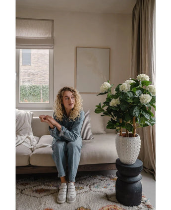 Een vrouw met krullend blond haar in spijkerstof en witte sneakers zit op een beige bank in een stijlvolle woonkamer met de Kunstplant Hortensia UV 85 cm wit, een neutrale inrichting en een raam dat natuurlijk licht binnenlaat.