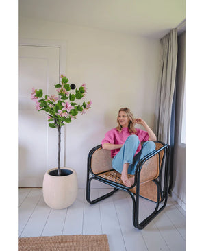 Een vrouw in een roze trui en blauwe spijkerbroek zit op blote voeten op een rotan stoel naast de Kunstplant Judas 150 cm, een kunstboom met roze bloemen, in een lichte, moderne kamer met witte muren en vloeren.