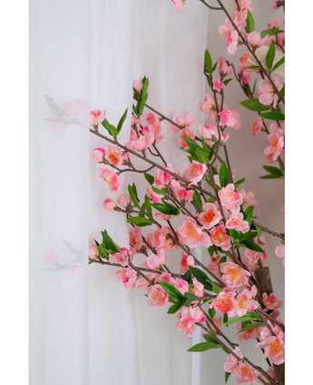 De Kunstplant Kersenbloesem 120 cm roze, met roze kersenbloesem en groene bladeren, staat naast een doorschijnend wit gordijn, de bloemen zacht zichtbaar door de doorschijnende stof.