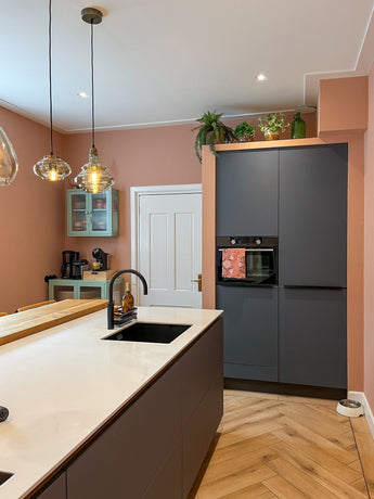 Cucina moderna con pareti color pesca, armadi grigio scuro, ripiani bianchi, lampade a sospensione e motivo a spina di pesce. Una pianta artificiale di Monstera da 18 cm e bottiglie di vetro sopra i mobili danno un tocco di freschezza.