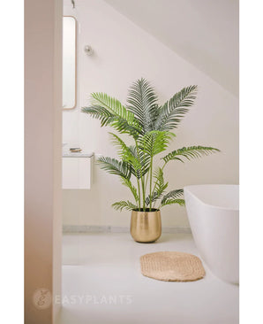 In een minimalistische badkamer met witte muren en armaturen staat de Kunstplant Phoenix 160 cm in een gouden plantenbak bij het bad voor een luxe uitstraling, aangevuld met een rond beige geweven vloerkleed op de vloer.