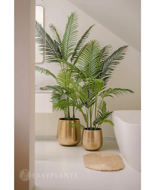 Una pianta artificiale Phoenix da 180 cm con foglie verdi lussureggianti in un vaso dorato lucido si trova accanto a una vasca bianca e un tappetino intrecciato beige in un bagno moderno e minimalista, completato da uno specchio e una finestra per un aspetto sereno.
