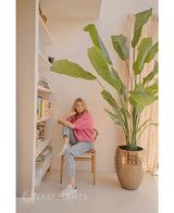 Een vrouw in een roze trui en spijkerbroek zit op een houten stoel naast de Kunstplant Strelitzia Nicolai Deluxe 235 cm; boekenplanken en transparante gordijnen versterken het zacht verlichte interieur.