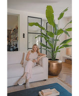 Een vrouw in lichte kleding zit met een mok op een beige bank. Naast haar brengt de Kunstplant Strelitzia Nicolai Deluxe 235 cm weelderig groen in de stijlvolle, moderne woonkamer met neutrale inrichting.