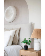 Een gezellige slaapkamer heeft een bed met neutraal beddengoed, een licht houten nachtkastje met de Kunstplant Watermeloen 29 cm en een witte lamp met een rieten kap. Boven het bed hangt een witte wandsculptuur met textuur.