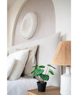 Een keurig opgemaakt bed met witte kussens en beddengoed staat naast een houten nachtkastje met de Kunstplant Watermeloen 29 cm en een lamp met een geweven kap; boven het hoofdeinde hangt een wandsculptuur met witte textuur.