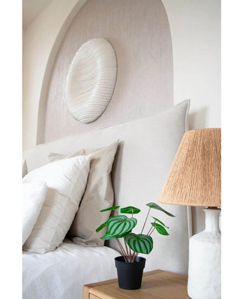 Een keurig opgemaakt bed met witte kussens en beddengoed staat naast een houten nachtkastje met de Kunstplant Watermeloen 29 cm en een lamp met een geweven kap; boven het hoofdeinde hangt een wandsculptuur met witte textuur.