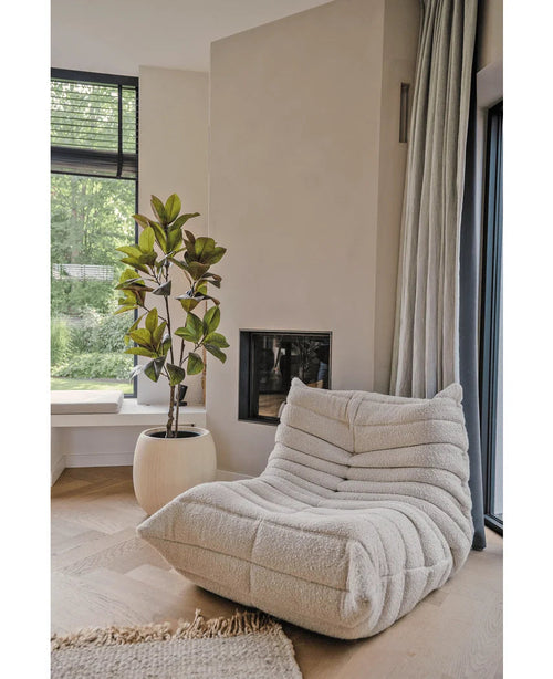 Un'accogliente poltrona lounge color crema strutturata si trova accanto a un camino moderno e un'alta finestra. Accanto c'è l'albero artificiale di Yulania 150 cm in un vaso rotondo bianco. I pavimenti in legno chiaro e la luce solare morbida creano un'atmosfera calma e invitante.