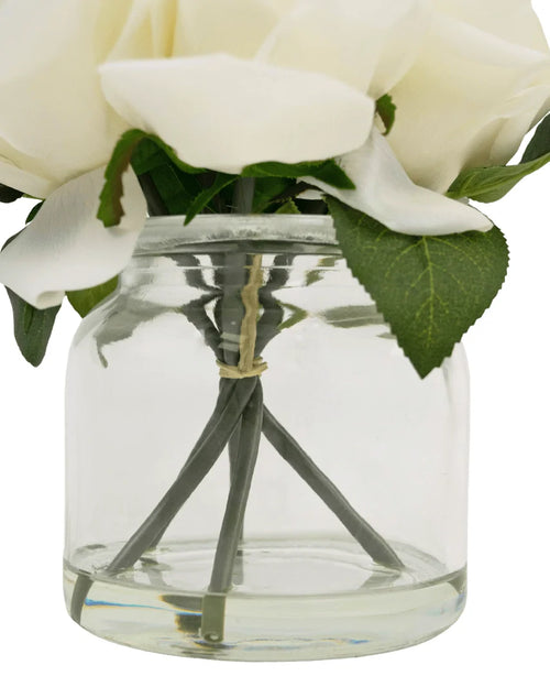Un primo piano del Bouquet di Rose Artificiali Real Touch White 18cm: fiori artificiali bianchi con foglie verdi in vasetto di vetro trasparente, steli a vista tenuti insieme da un elastico.