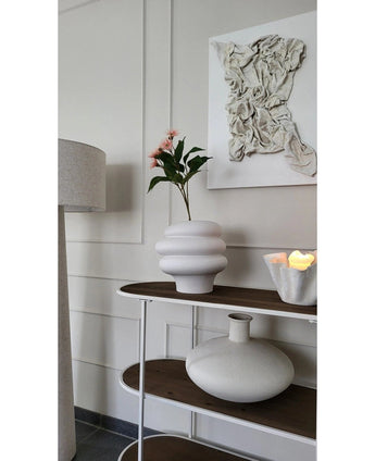 Een modern interieur met een wit wandkunstwerk met textuur, een witte sculpturale vaas met Kunsttak Bloeiende Eucalyptus 62 cm oud roze, een decoratieve kaars en een ronde keramische vaas op een houten plank naast een staande lamp.