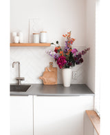 Una cucina moderna bianca con piano di lavoro grigio, un lavello, un tagliere in legno e un vaso bianco pieno di fiore artificiale di ortensia da 55 cm rosa scuro. Tazze e pentole bianche stanno su uno scaffale di legno sopra il bancone.