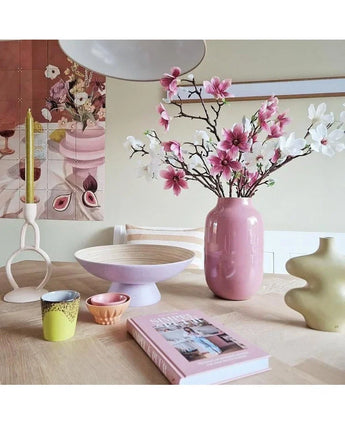 Een moderne kamer heeft een gedekte tafel met Kunsttak Magnolia kleinbloemig 89 cm roze in een roze vaas, pastelkleurige kandelaars, schalen, kopjes, een groene sculpturale vaas, een roze boek en bloemenkunstwerk aan de muur.