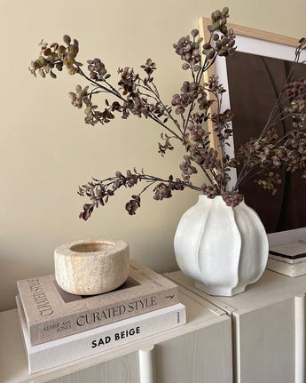 Op een lichte kast staat een witte vaas gevuld met de Kunsttak Eucalyptus bes 110 cm bruin, naast een stapel boeken en een stenen kandelaar, met op de achtergrond een ingelijste foto.