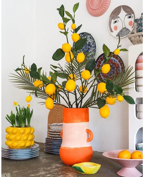Una stanza luminosa con un vaso alto pieno di limoni e foglie verdi, in cui il sorprendente Kunsttak Lemon 104 cm giallo occupa un posto di rilievo nella disposizione, circondato da ciotole in ceramica colorate, una ciotola di limoni e piatti decorativi sulla parete, creando un look allegro ed eclettico.