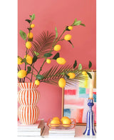 Tegen een koraalroze muur staan een gestreepte vaas gevuld met Kunsttak Lemon-takken van 104 cm in geel, een kleurrijk schilderij, een stapel boeken, een blauwe kandelaar met een kaars en een dienblad met citroenen.