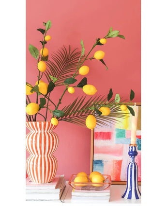 Tegen een koraalroze muur staan een gestreepte vaas gevuld met Kunsttak Lemon-takken van 104 cm in geel, een kleurrijk schilderij, een stapel boeken, een blauwe kandelaar met een kaars en een dienblad met citroenen.
