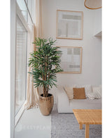 Een Lady Kunstpalm 180 cm is geplaatst bij een groot raam in een lichte, moderne woonkamer met een witte bank, beige kussens, geweven tafel en abstracte kunst. Het voegt een gezellige minimalistische stijl toe met zijn neutrale tinten en groene kunstplant charme.