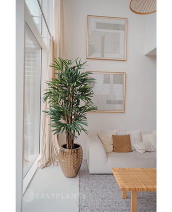 Een Lady Kunstpalm 180 cm is geplaatst bij een groot raam in een lichte, moderne woonkamer met een witte bank, beige kussens, geweven tafel en abstracte kunst. Het voegt een gezellige minimalistische stijl toe met zijn neutrale tinten en groene kunstplant charme.