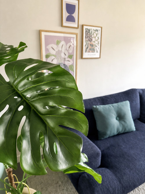 Nella parte anteriore è ben visibile una pianta artificiale Monstera di 120 cm. Dietro c'è un divano blu scuro con un cuscino azzurro contro una parete beige con tre opere d'arte incorniciate.